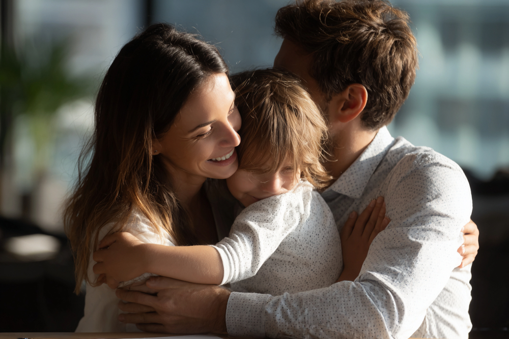 Two parents embracing their young child indoors with a city view through the window behind them
