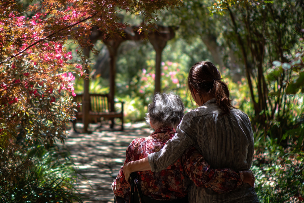 Adult daughter with her arm around an older woman in a flowering garden, representing guardianship for an aging loved one