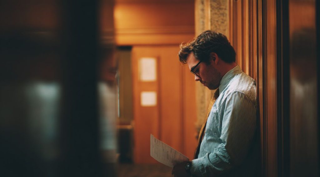 Man in a shirt and tie reading a document in a wood-panelled courthouse corridor