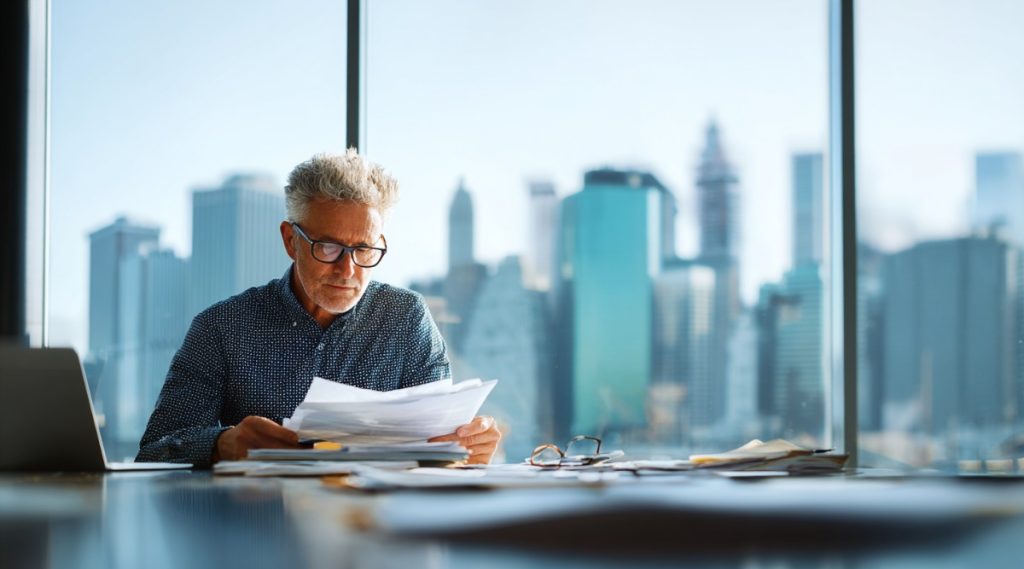 Older man reviewing papers and eyeglasses at a glass table before a city skyline, on valuing closely held businesses
