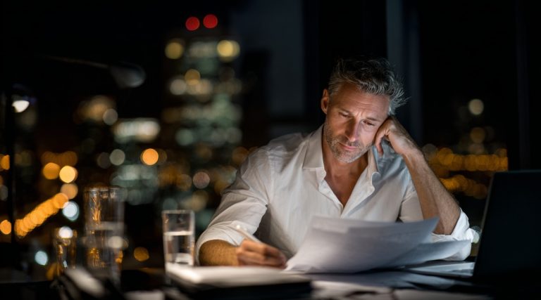 Man alone at his desk at night reviewing papers with city lights behind him, on whether filing first matters